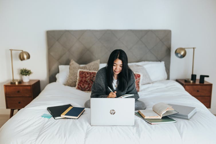 A woman sits on her bed working on her laptop with books and notebooks surrounding her.