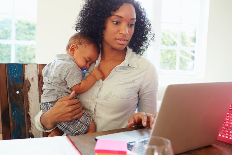 A woman holds a baby while looking at her laptop screen.