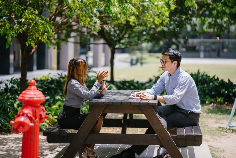 A man and woman sit outside at a picnic table talking in an urban setting, with a red fire hydrant in the foreground. The man takes notes.