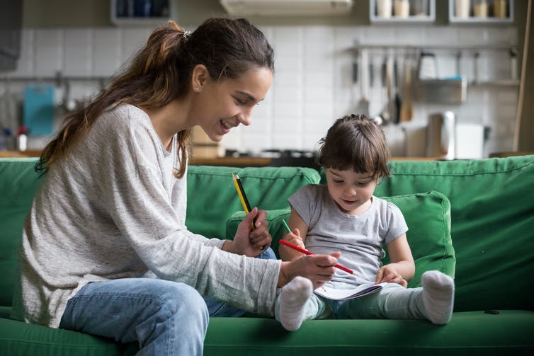 A teenage girl and a preschool aged girl colour with pencil crayons on a sofa.