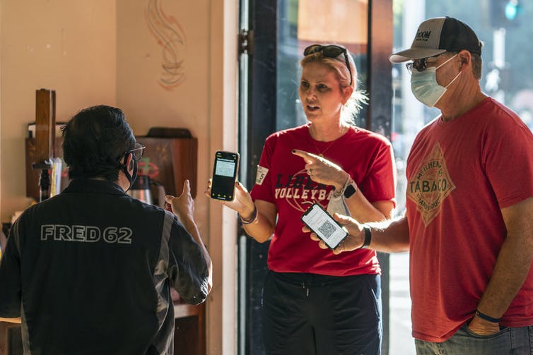 Two customers – one wearing a mask, one not – hold up their phones to a waiter wearing a mask to show their vaccination status