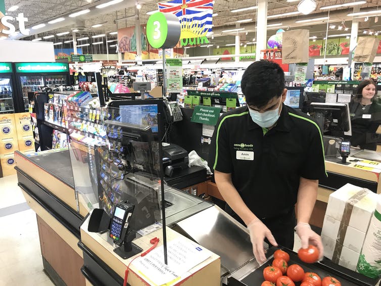 A grocery store cashier, wearing a mask and gloves, works behind a Plexiglas barrier.