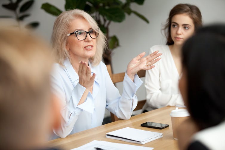 Women in glasses speaking to other women at a table in a meeting room