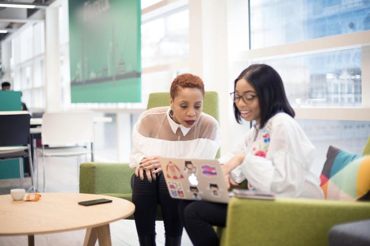 Two women are seen seated in a room looking at a laptop.