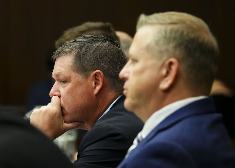 Two men, one with his hand against his jaw, sit in a hearing room.