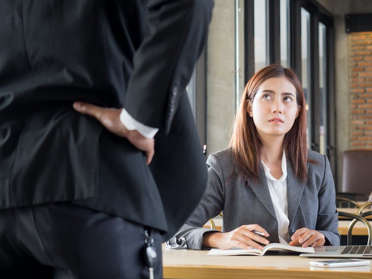 A woman in a blazer looks up nervously at a man with his hands on his hips. The man has his back to us and is standing very close to the camera.