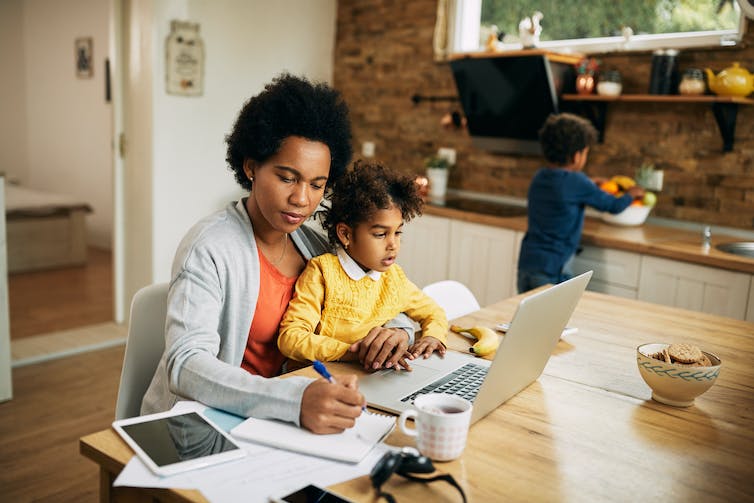 A woman sits at a kitchen table behind a laptop with a young girl on her lap. A young boy is seen picking fruit out of a bowl in the background.