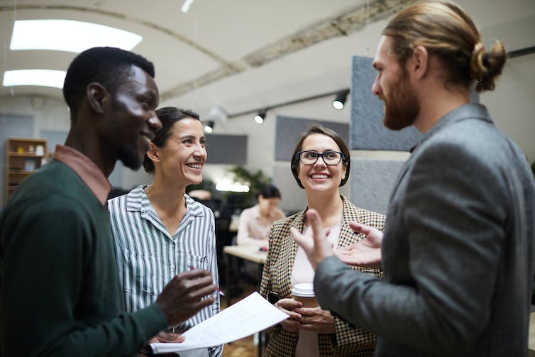 A group of diverse people chat together in front of people working in cubicles