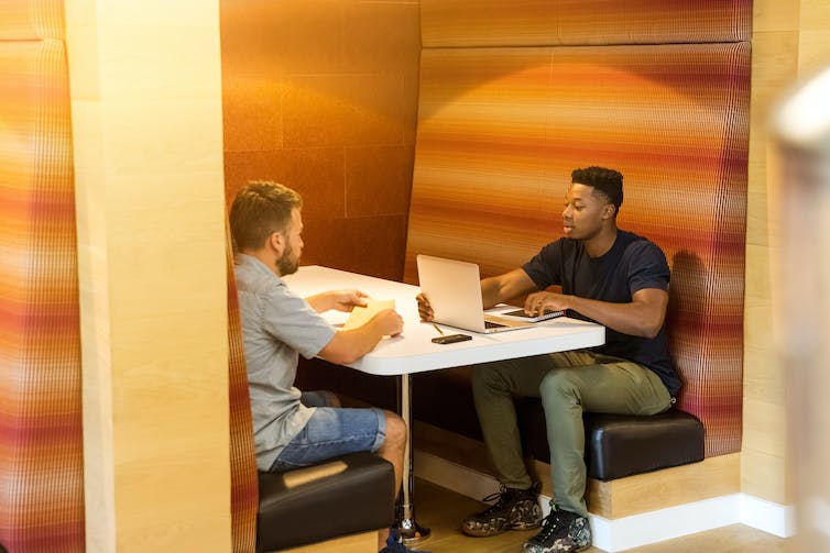 two men sit at a desk talking.