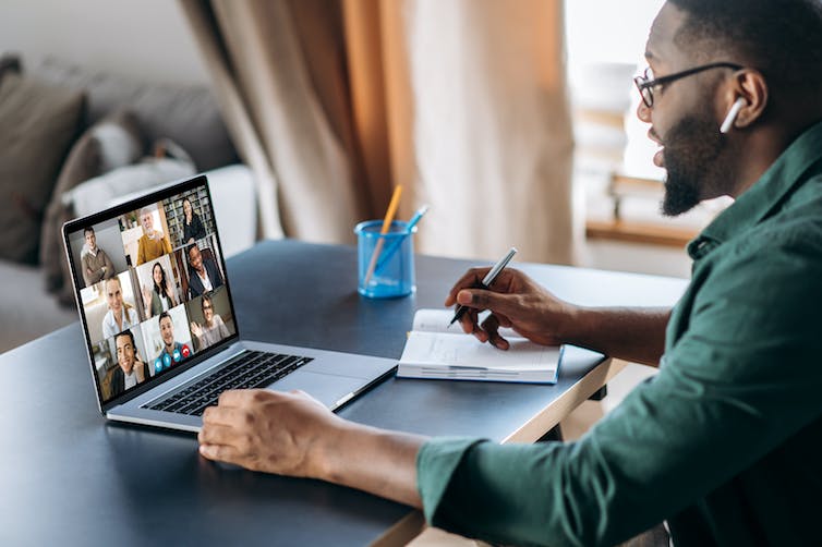 A man, wearing wireless ear buds and sitting at a desk, speaks on a video call