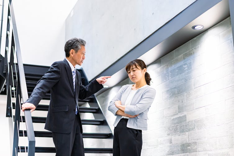 An older man in a business suit points and talks sternly to a younger woman who is also in business attire