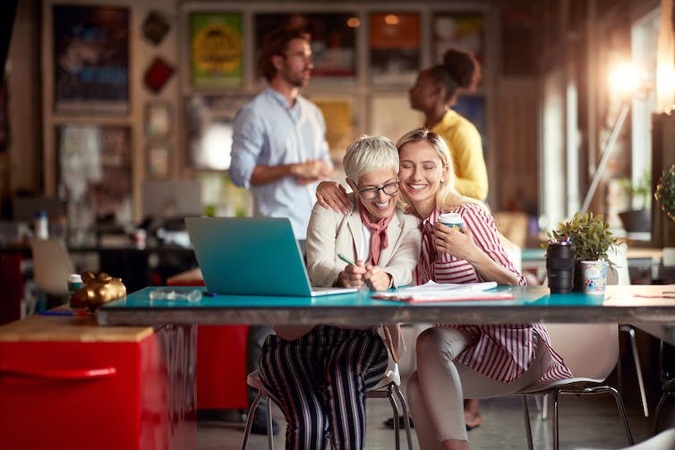 A young woman at the desk in the office hugs an older female colleague.