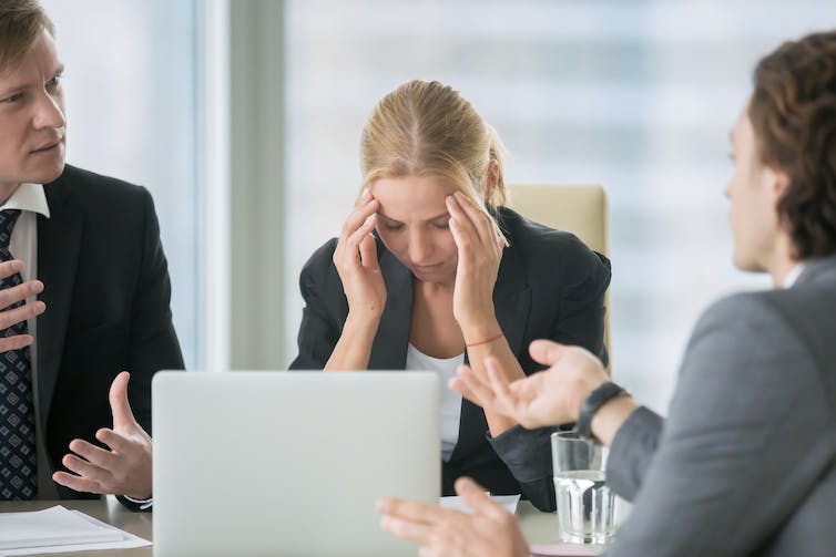 A woman sits with her head in her hands as two men speak animatedly at her