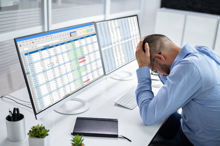 Employee with head in hands in front of computer screens.