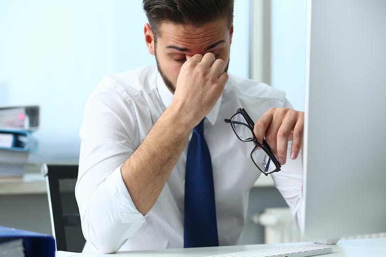 A man in a white dress shirt and tie pinching the bridge of his nose. He is seated at a desk in front of a computer screen.