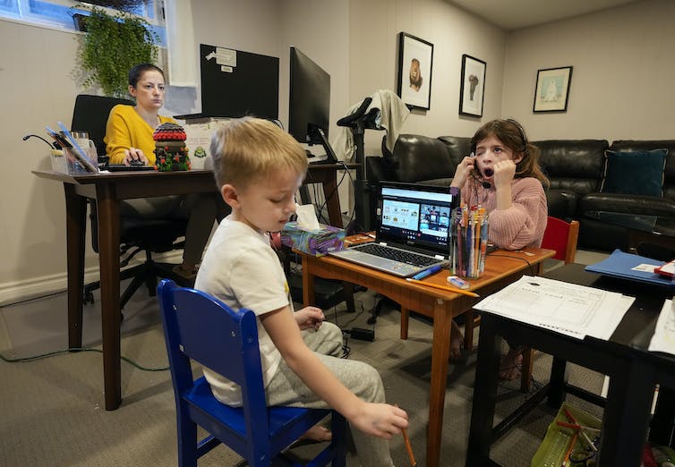 Two children sit at laptops while their mother sits at a desk looking at her own laptop.