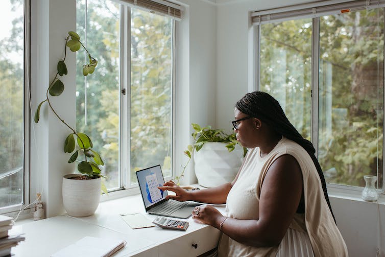 A woman working at a laptop at a desk in a room with large windows.