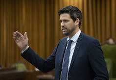 A bearded man in a dark blue suit gestures as he speaks.