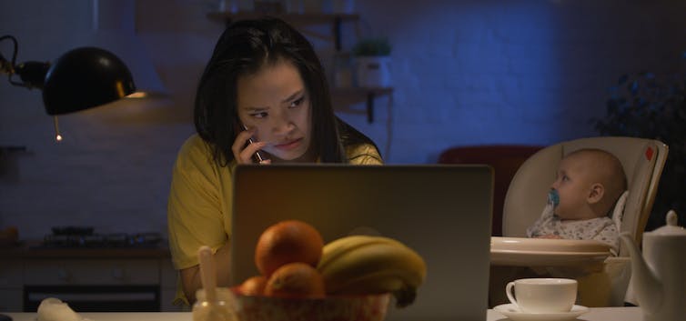 A woman working on a laptop with a baby beside her in a high chair