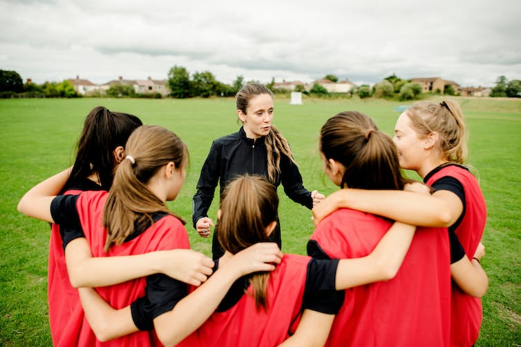 A woman coach in a black, long-sleeved shirt speaks to a group of young girls wearing red pinnies