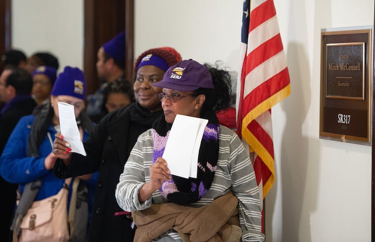 A row of people stand in a hall and wear purple hats and hold up white papers.