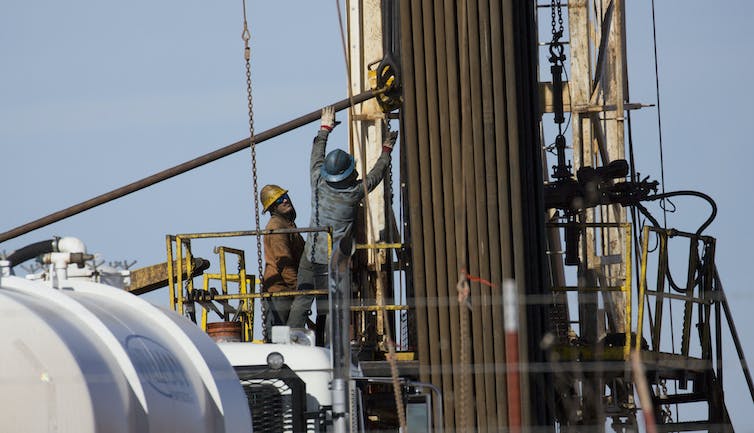 Workers in hard hats reach for pipes in a tall stand of pipes at a finishing well in Oklahoma.