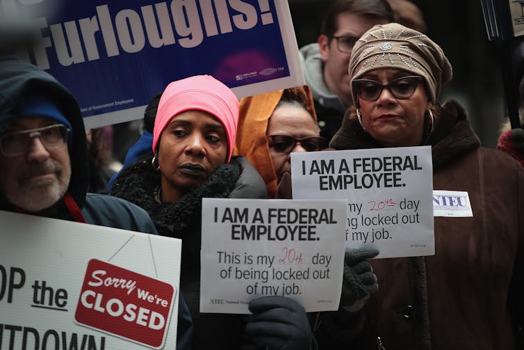 A group of people wear hats and warm clothing and hold up signs that say 'I am a federal employee' and 'Sorry, we're closed.'