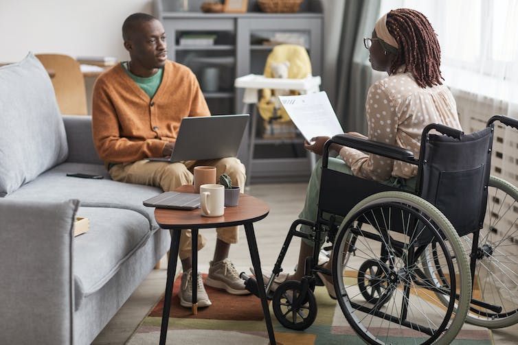 A woman in a wheechair speaks with a man seated on a sofa. Both are using laptops.