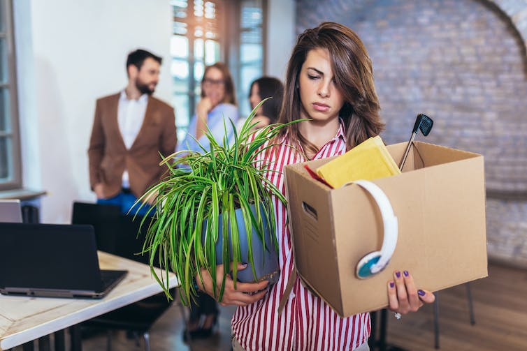 Woman with box of belongings and plant, looking annoyed, people talking in the background.