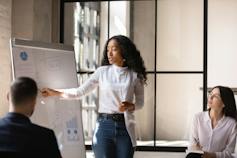 A young Black women points to a whitebaord while giving a presentation to a table of people.