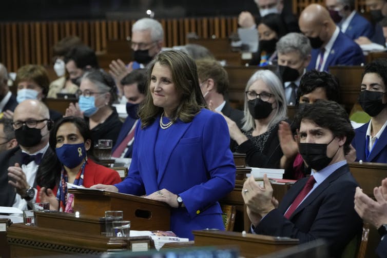 A woman in a suit standing behind a podium. Behind her, a crowd of people in masks and business attire sit and watch her.