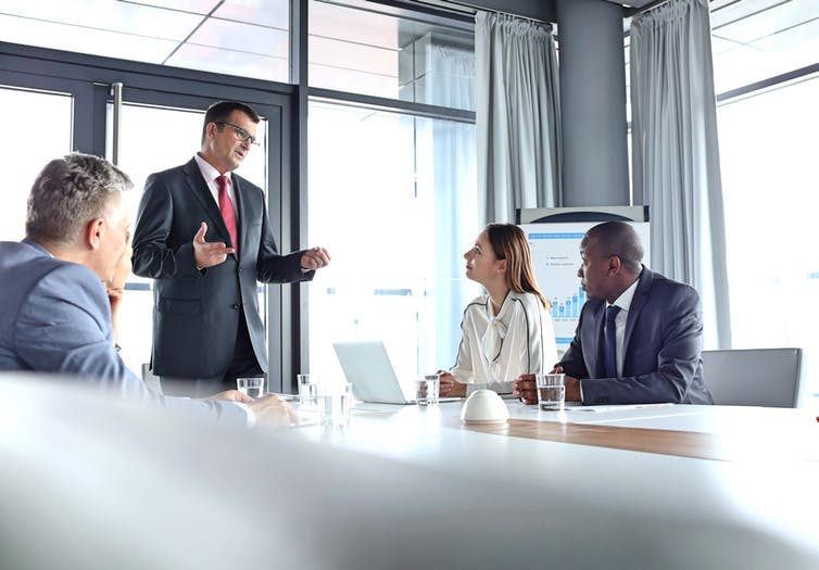 A business man stands at a table in a meeting room talking to a woman and a Black man.